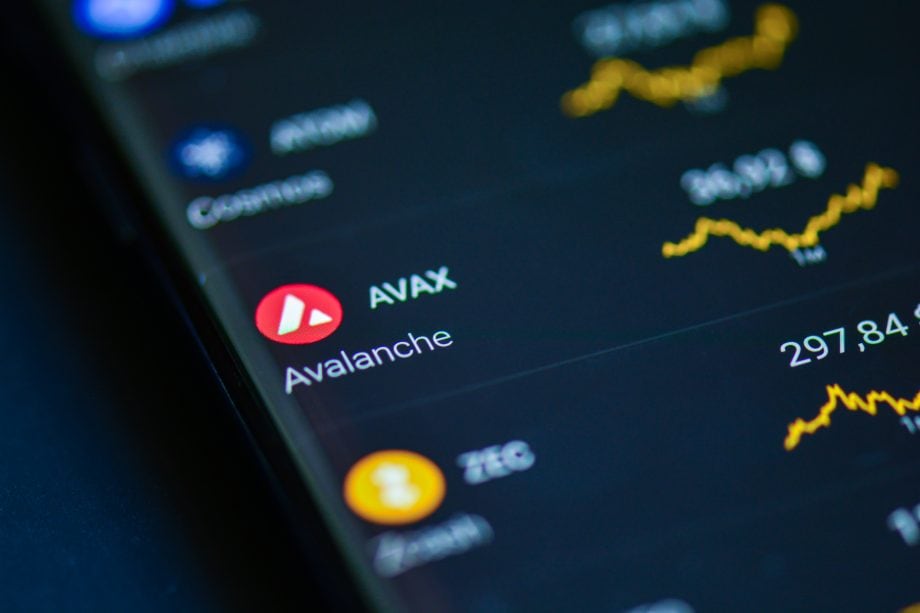 Das Bild zeigt die Kryptowährung Avalanche (AVAX) auf dem Display eines Smartphones oder Tablets. Im Fokus stehen das rote Avalanche-Logo und die Bezeichnung "AVAX". Im Hintergrund sind unscharfe Kurscharts und weitere Kryptowährungen zu erkennen, darunter Bitcoin. Die Aufnahme vermittelt den Eindruck einer Handels- oder Portfolio-App.