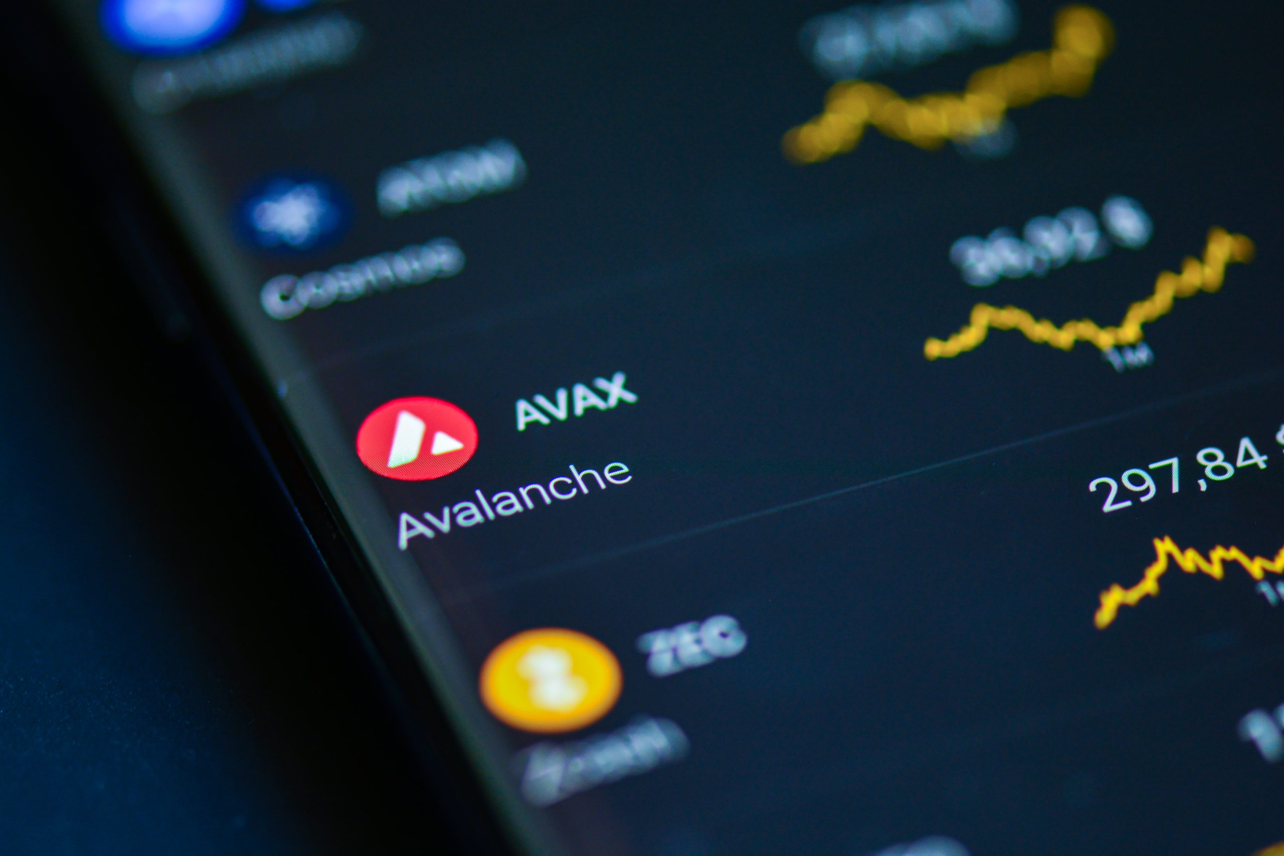 Das Bild zeigt die Kryptowährung Avalanche (AVAX) auf dem Display eines Smartphones oder Tablets. Im Fokus stehen das rote Avalanche-Logo und die Bezeichnung "AVAX". Im Hintergrund sind unscharfe Kurscharts und weitere Kryptowährungen zu erkennen, darunter Bitcoin. Die Aufnahme vermittelt den Eindruck einer Handels- oder Portfolio-App.