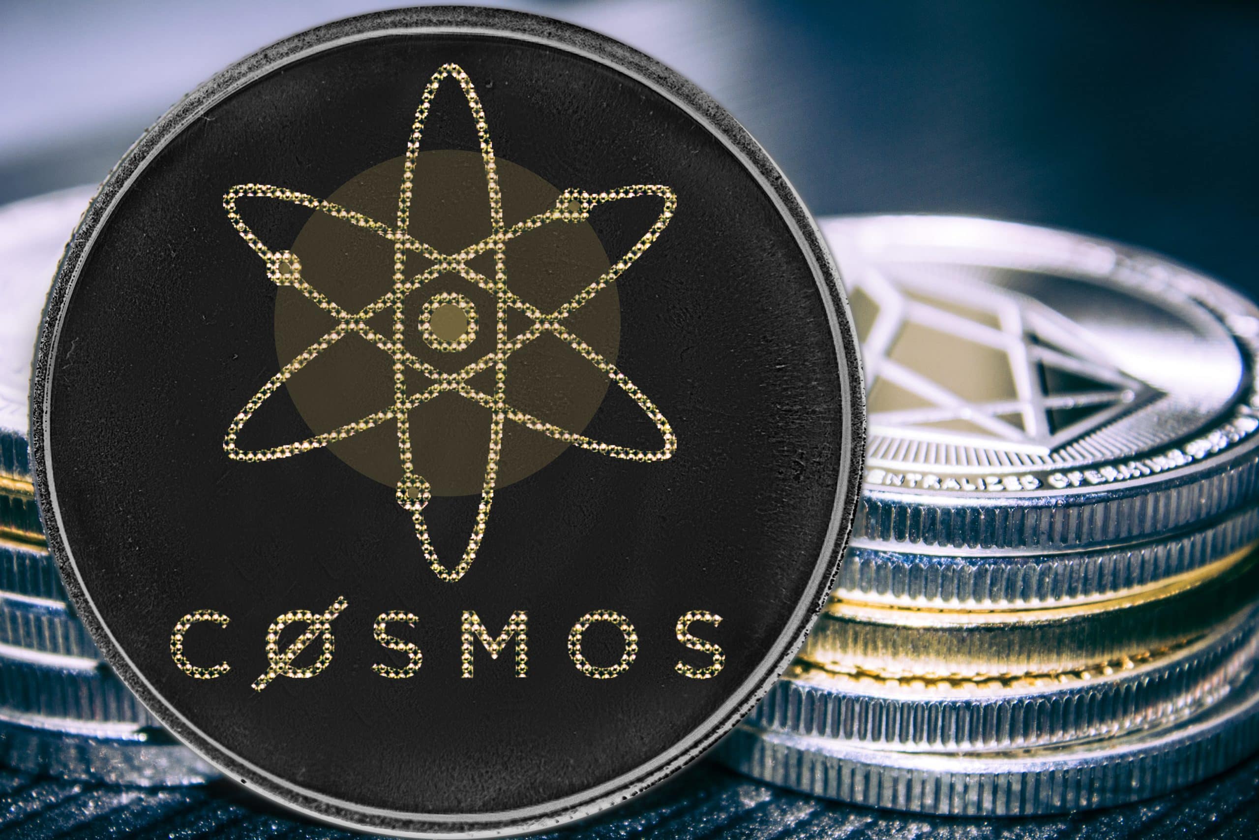 Das Bild zeigt eine Cosmos-(ATOM)-Münze im Vordergrund, die auf mehreren übereinandergestapelten Krypto-Coins liegt. Im Zentrum der schwarzen Münze ist das charakteristische Atom-Symbol von Cosmos abgebildet. Im Hintergrund sind unscharf weitere Kryptowährungen zu erkennen, darunter eine Ethereum-Münze.