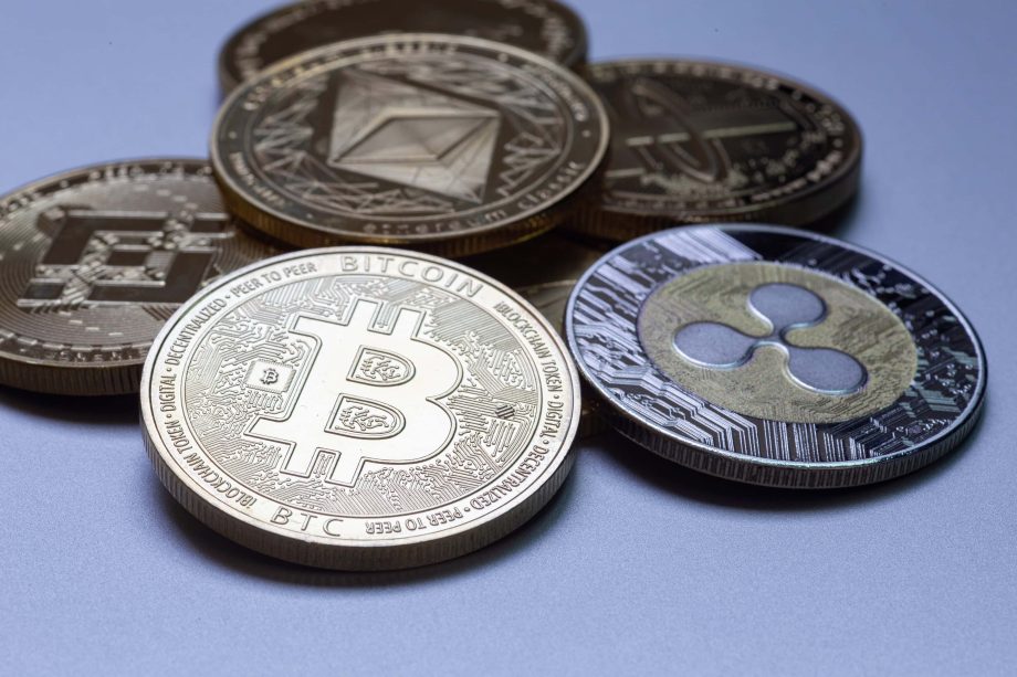 Mehrere physische Münzen, die verschiedene Kryptowährungen repräsentieren, darunter Bitcoin, Ethereum und Ripple, sind auf einer glatten, grauen Oberfläche angeordnet.