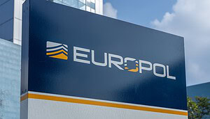 Das Bild zeigt ein großes Europol-Schild vor einem modernen Bürogebäude mit Glasfassade. Das blau-weiße Logo der europäischen Polizeibehörde ist zentral im Vordergrund platziert und dominiert die Aufnahme. Im Hintergrund sind weitere Gebäudeteile sowie ein teilweise bewölkter Himmel zu erkennen.
