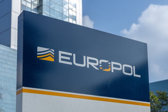 Das Bild zeigt ein großes Europol-Schild vor einem modernen Bürogebäude mit Glasfassade. Das blau-weiße Logo der europäischen Polizeibehörde ist zentral im Vordergrund platziert und dominiert die Aufnahme. Im Hintergrund sind weitere Gebäudeteile sowie ein teilweise bewölkter Himmel zu erkennen.