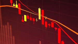 Das Bild zeigt einen stark fallenden Kurschart auf einem Bildschirm. Dominant sind rote Candlesticks, die einen anhaltenden Abwärtstrend visualisieren. Zwei gleitende Durchschnittslinien verlaufen ebenfalls nach unten und unterstreichen die negative Kursentwicklung. Der Hintergrund ist in dunklen Rot- und Schwarztönen gehalten, wodurch die roten Kurskerzen besonders hervorstechen.