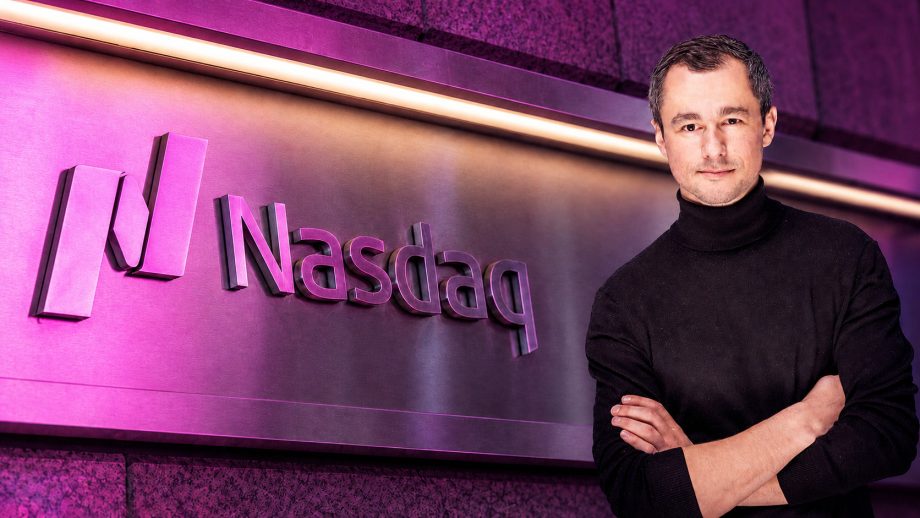 Das Logo der Nasdaq wird violett angeleuchtet. Rechtbündig steht ein Mann mit einem schwarzen Rollkragenpullover.