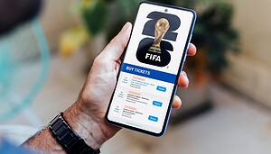 Das Bild zeigt eine Person, die ein Smartphone in der Hand hält. Auf dem Display ist eine Ticketseite zur FIFA-Weltmeisterschaft zu sehen, inklusive FIFA-Logo, WM-Pokal und mehreren Ticketangeboten. Der Fokus liegt auf dem digitalen Ticketkauf, während der Hintergrund unscharf bleibt.