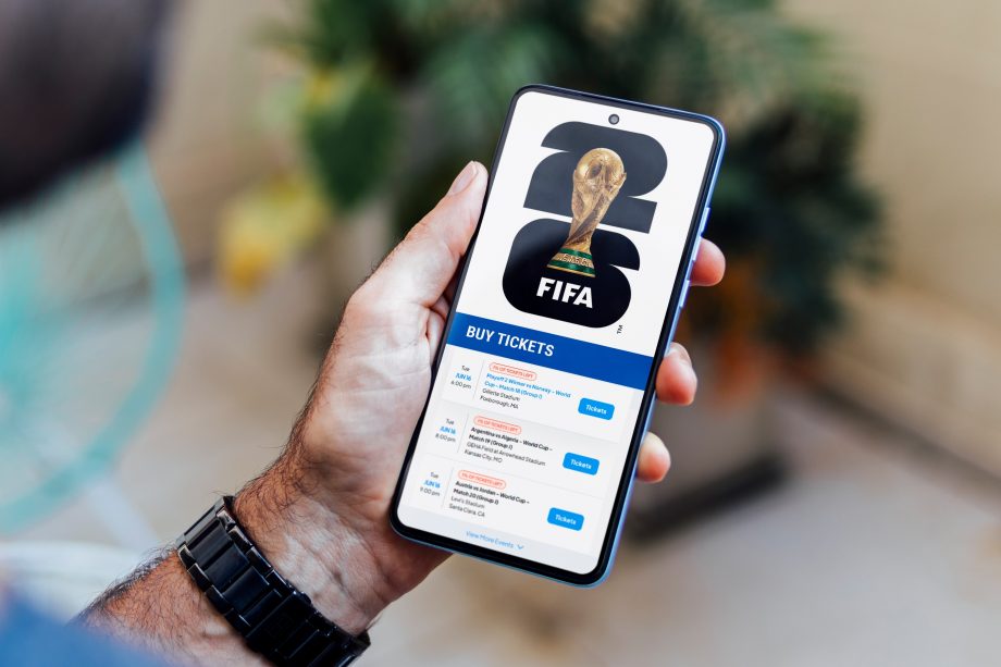 Das Bild zeigt eine Person, die ein Smartphone in der Hand hält. Auf dem Display ist eine Ticketseite zur FIFA-Weltmeisterschaft zu sehen, inklusive FIFA-Logo, WM-Pokal und mehreren Ticketangeboten. Der Fokus liegt auf dem digitalen Ticketkauf, während der Hintergrund unscharf bleibt.