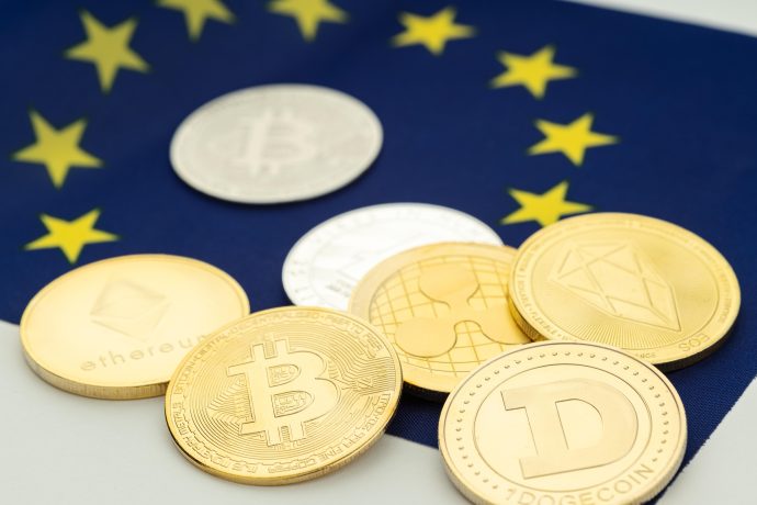 Das Bild zeigt mehrere physische Kryptowährungs-Token auf einer Flagge der Europäischen Union. Zu erkennen sind unter anderem Bitcoin, Ethereum, XRP, EOS und Dogecoin. Die EU-Sterne im Hintergrund sind leicht unscharf dargestellt, während die Krypto-Coins im Vordergrund liegen.