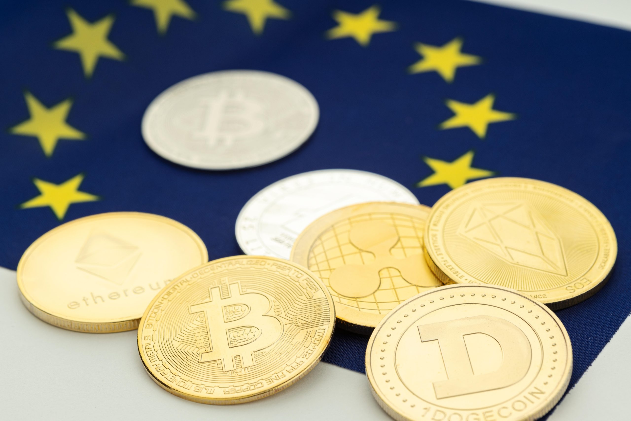 Das Bild zeigt mehrere physische Kryptowährungs-Token auf einer Flagge der Europäischen Union. Zu erkennen sind unter anderem Bitcoin, Ethereum, XRP, EOS und Dogecoin. Die EU-Sterne im Hintergrund sind leicht unscharf dargestellt, während die Krypto-Coins im Vordergrund liegen.