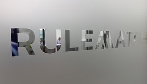 Das Bild zeigt den Schriftzug „RULEMATCH“, der in eine matte Glas- oder Milchglasfläche eingelassen ist. Die Buchstaben sind ausgespart und geben den Blick auf den dahinterliegenden Innenraum frei. Im Hintergrund ist unscharf eine Person in einem modernen Büro oder Besprechungsraum zu erkennen. Die Aufnahme ist in neutralen Grau- und Weißtönen gehalten und vermittelt den Eindruck eines Unternehmenssitzes oder Bürostandorts.