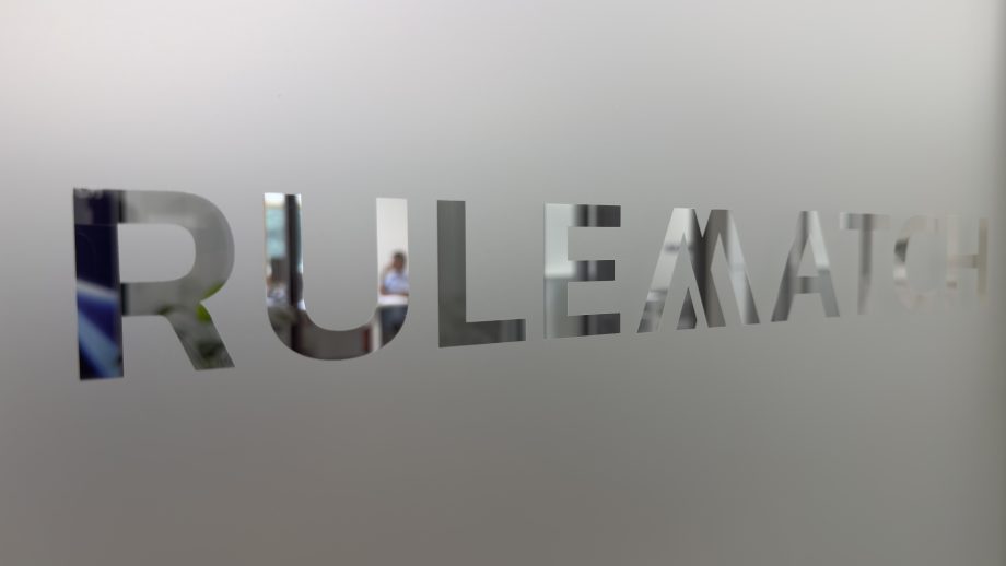 Das Bild zeigt den Schriftzug „RULEMATCH“, der in eine matte Glas- oder Milchglasfläche eingelassen ist. Die Buchstaben sind ausgespart und geben den Blick auf den dahinterliegenden Innenraum frei. Im Hintergrund ist unscharf eine Person in einem modernen Büro oder Besprechungsraum zu erkennen. Die Aufnahme ist in neutralen Grau- und Weißtönen gehalten und vermittelt den Eindruck eines Unternehmenssitzes oder Bürostandorts.