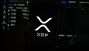 Das Bild zeigt ein Smartphone mit dem XRP-Logo auf schwarzem Hintergrund. Im Hintergrund sind unscharfe Monitore oder Dashboards zu erkennen, die an Handels-, Analyse- oder Netzwerkinfrastruktur erinnern. Die dunkle Farbgebung und der Fokus auf das XRP-Symbol vermitteln einen technischen und institutionellen Eindruck.