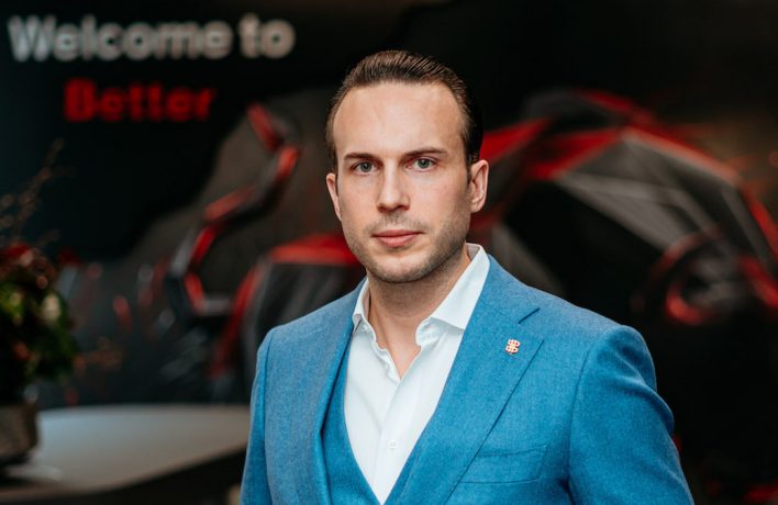 Ein Mann mit blauem Anzug steht vor einem Hintergrund in den Farben der Bitcoin Suisse