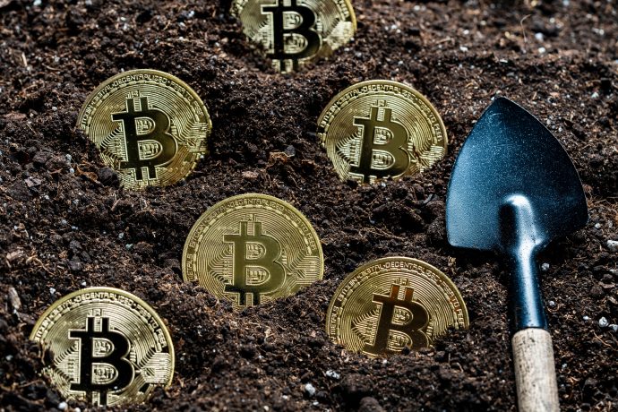 Eine kleine Schaufel liegt neben mehreren in Erde eingegrabenen Bitcoin-Münzen.