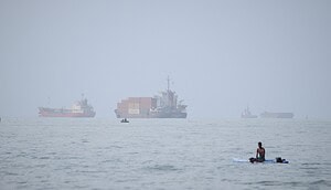 Ein Mann sitzt auf einem Surfbrett, während ein Containerschiff und andere Handelsschiffe vor Bandar Abbas im Iran in der Straße von Hormus vor Anker liegen.