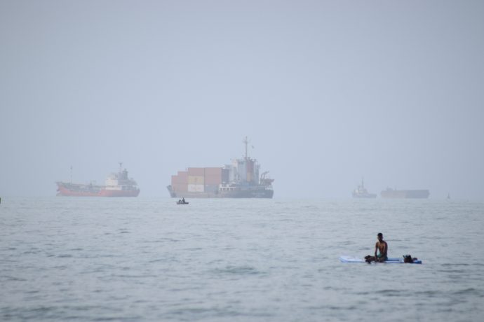 Ein Mann sitzt auf einem Surfbrett, während ein Containerschiff und andere Handelsschiffe vor Bandar Abbas im Iran in der Straße von Hormus vor Anker liegen.