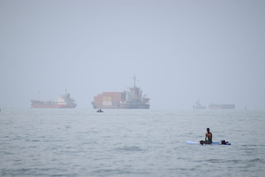 Ein Mann sitzt auf einem Surfbrett, während ein Containerschiff und andere Handelsschiffe vor Bandar Abbas im Iran in der Straße von Hormus vor Anker liegen.