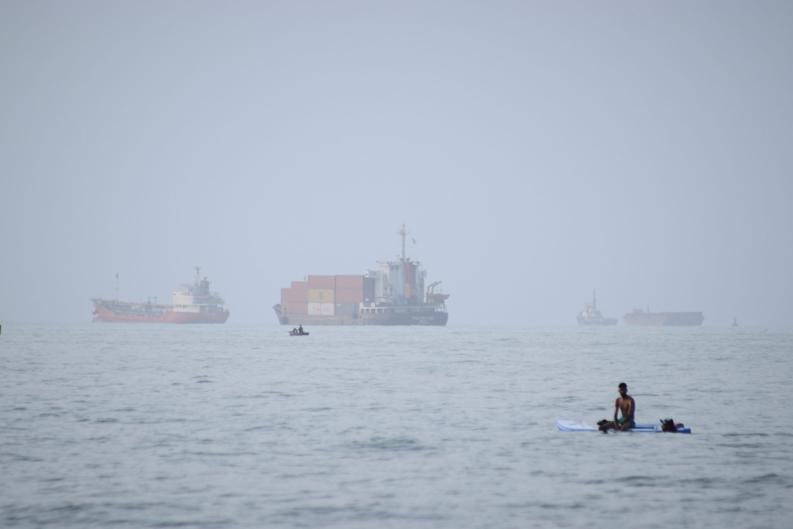 Ein Mann sitzt auf einem Surfbrett, während ein Containerschiff und andere Handelsschiffe vor Bandar Abbas im Iran in der Straße von Hormus vor Anker liegen.
