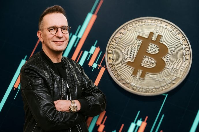 Investmentpunk Gerald Hörhan steht mit verschränkten Armen vor einem Bitcoin-Kurschart und einer großen Bitcoin-Münze.