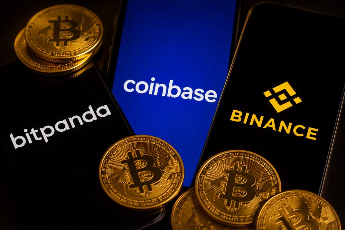 Mehrere Smartphones mit den Logos der Krypto-Börsen Bitpanda, Coinbase und Binance stehen nebeneinander. Im Vordergrund liegen mehrere goldfarbene Bitcoin-Münzen. Das Bild symbolisiert den Wettbewerb und die Präsenz großer Krypto-Handelsplattformen.