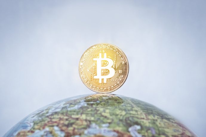 Man sieht einen Globus auf dem eine Bitcoin-Münze steht vor blauem Hintergrund.