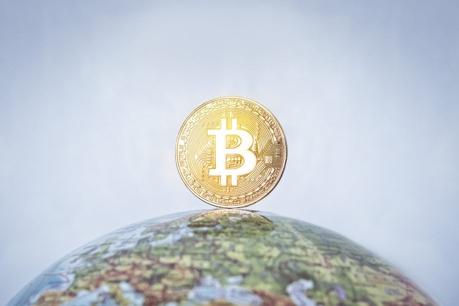 Man sieht einen Globus auf dem eine Bitcoin-Münze steht vor blauem Hintergrund.