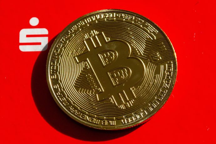 Das Bild zeigt eine Bitcoin-Münze vor dem Logo der Sparkassen
