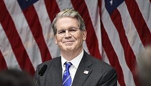 Ein Mann in Anzug und Krawatte (US-Finanzminister Scott Bessent) steht vor mehreren amerikanischen Flaggen und lächelt von einem Podium mit einem Mikrofon, als der US-Finanzminister neue Bitcoin-Regelungen ankündigt.