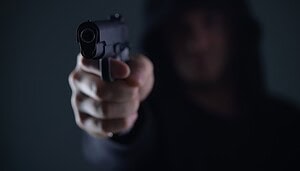 Eine Person mit einem dunklen Kapuzenpullover richtet eine Pistole auf die Kamera, was auf einen Krypto-Raub hindeutet. Die Waffe ist scharf abgebildet, während das Gesicht im Hintergrund unscharf bleibt.