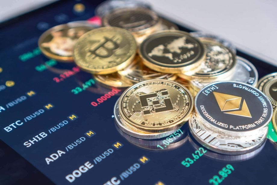 Physische Darstellungen verschiedener Kryptowährungen, darunter Bitcoin, Ethereum und Binance Coin, ruhen auf einem digitalen Bildschirm, der die Marktdaten des Krypto-Marktes anzeigt.