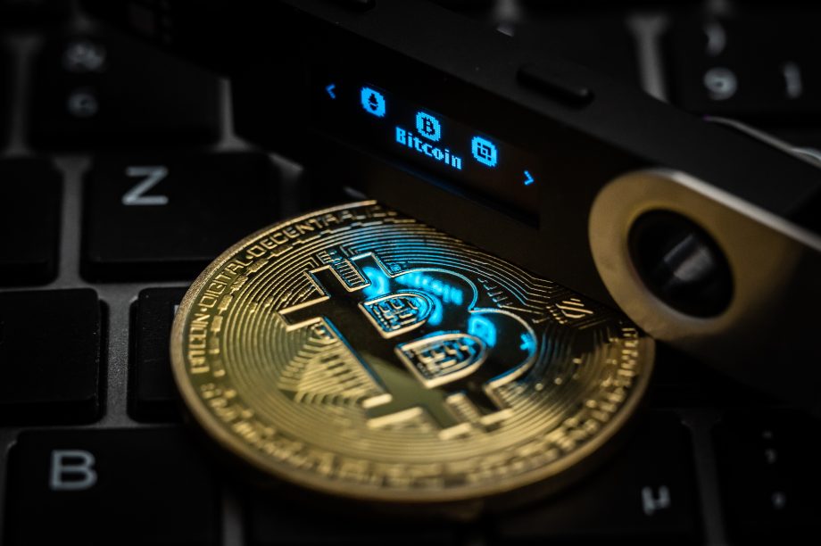 Der Schriftzug Bitcoin ist auf dem Display eines Ledger Nano S Hardware Wallets neben einer symbolischen „Bitcoin Münze“ zu sehen.