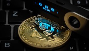 Der Schriftzug Bitcoin ist auf dem Display eines Ledger Nano S Hardware Wallets neben einer symbolischen „Bitcoin Münze“ zu sehen.