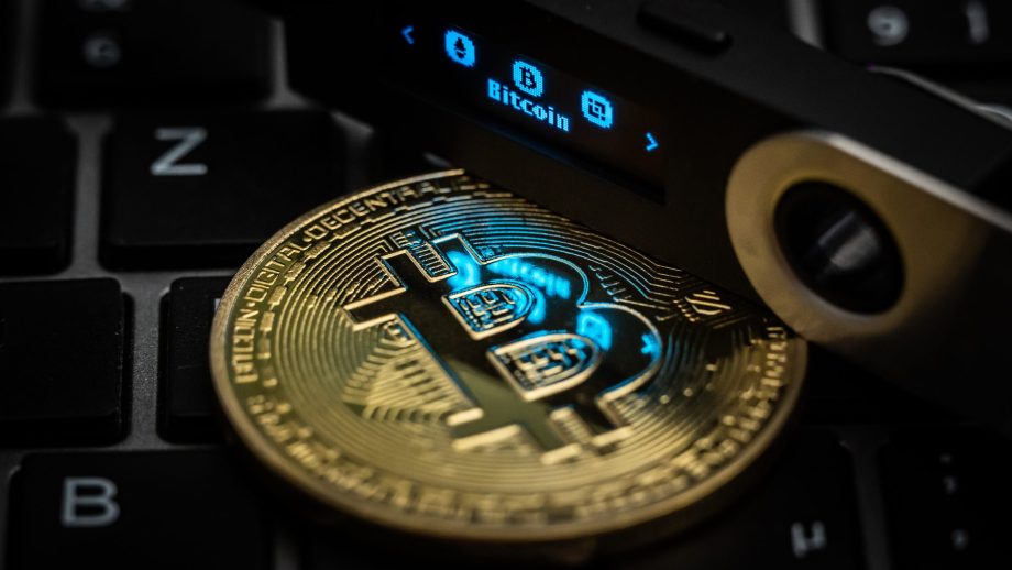Der Schriftzug Bitcoin ist auf dem Display eines Ledger Nano S Hardware Wallets neben einer symbolischen „Bitcoin Münze“ zu sehen.