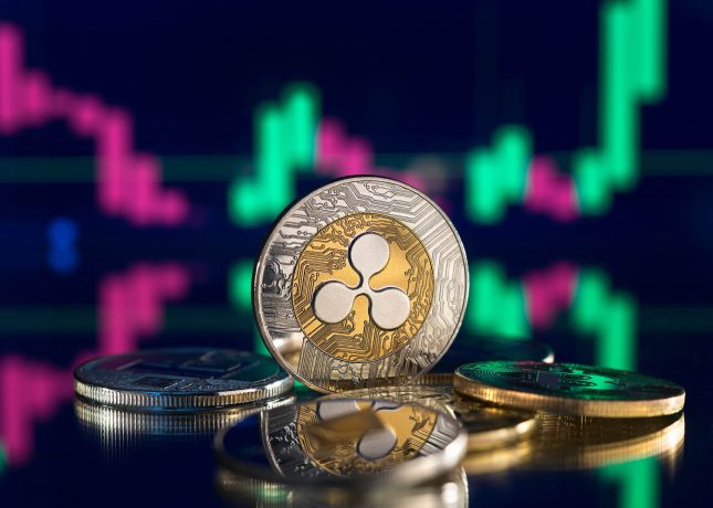 Eine Nahaufnahme von Ripple (XRP)-Kryptowährungsmünzen auf einer spiegelnden Oberfläche mit einem digitalen Preisdiagramm, das den XRP-Kurs und leuchtend grüne und rosafarbene Kerzen im Hintergrund zeigt.