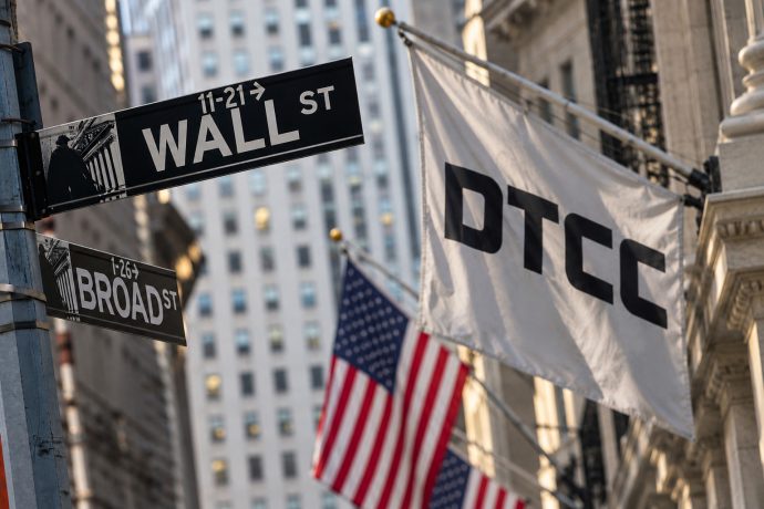 Wall-Street-Straßenschild in New York neben einer weißen Flagge mit DTCC-Logo und US-amerikanischen Flaggen im Hintergrund.