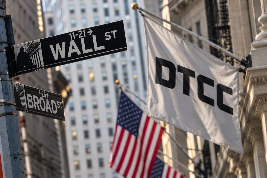 Wall-Street-Straßenschild in New York neben einer weißen Flagge mit DTCC-Logo und US-amerikanischen Flaggen im Hintergrund.
