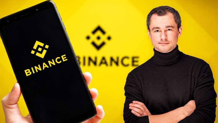 Binance Logo im Hintergrund und Binance Logo auf einem Smartphone links. Rechts steht ein Mann mit schwarzem Rollkragenpullover.
