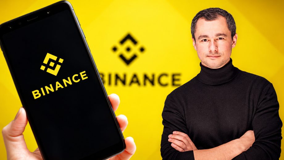 Binance Logo im Hintergrund und Binance Logo auf einem Smartphone links. Rechts steht ein Mann mit schwarzem Rollkragenpullover.