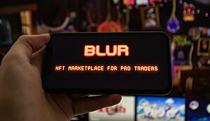 Eine Hand hält ein Smartphone, auf dessen Display das rot leuchtende Blur-Logo sowie der Schriftzug "NFT Marketplace for Pro Traders" zu sehen sind. Im unscharfen Hintergrund stehen mehrere Bildschirme mit bunten digitalen Motiven.
