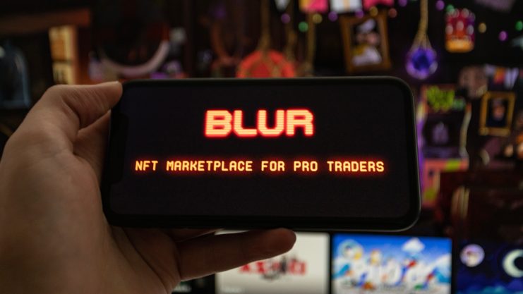 Eine Hand hält ein Smartphone, auf dessen Display das rot leuchtende Blur-Logo sowie der Schriftzug "NFT Marketplace for Pro Traders" zu sehen sind. Im unscharfen Hintergrund stehen mehrere Bildschirme mit bunten digitalen Motiven.