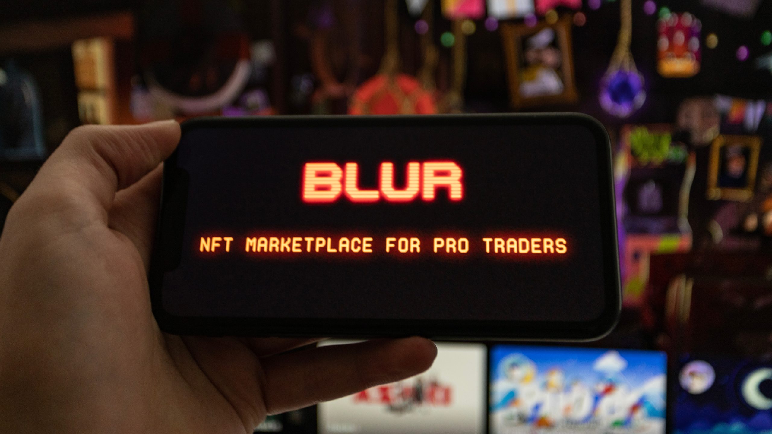 Eine Hand hält ein Smartphone, auf dessen Display das rot leuchtende Blur-Logo sowie der Schriftzug "NFT Marketplace for Pro Traders" zu sehen sind. Im unscharfen Hintergrund stehen mehrere Bildschirme mit bunten digitalen Motiven.
