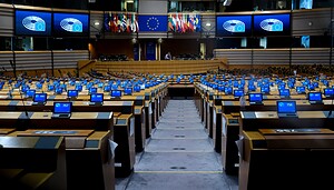 Das Bild zeigt den Plenarsaal des Europäischen Parlaments in Brüssel. Zu sehen sind die weitgehend leeren Sitzreihen der Abgeordneten. Auf mehreren Bildschirmen ist das Logo des Europäischen Parlaments vor blauem Hintergrund eingeblendet, im Zentrum hängen die EU-Flagge sowie die Flaggen der Mitgliedstaaten.
