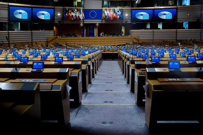 Das Bild zeigt den Plenarsaal des Europäischen Parlaments in Brüssel. Zu sehen sind die weitgehend leeren Sitzreihen der Abgeordneten. Auf mehreren Bildschirmen ist das Logo des Europäischen Parlaments vor blauem Hintergrund eingeblendet, im Zentrum hängen die EU-Flagge sowie die Flaggen der Mitgliedstaaten.