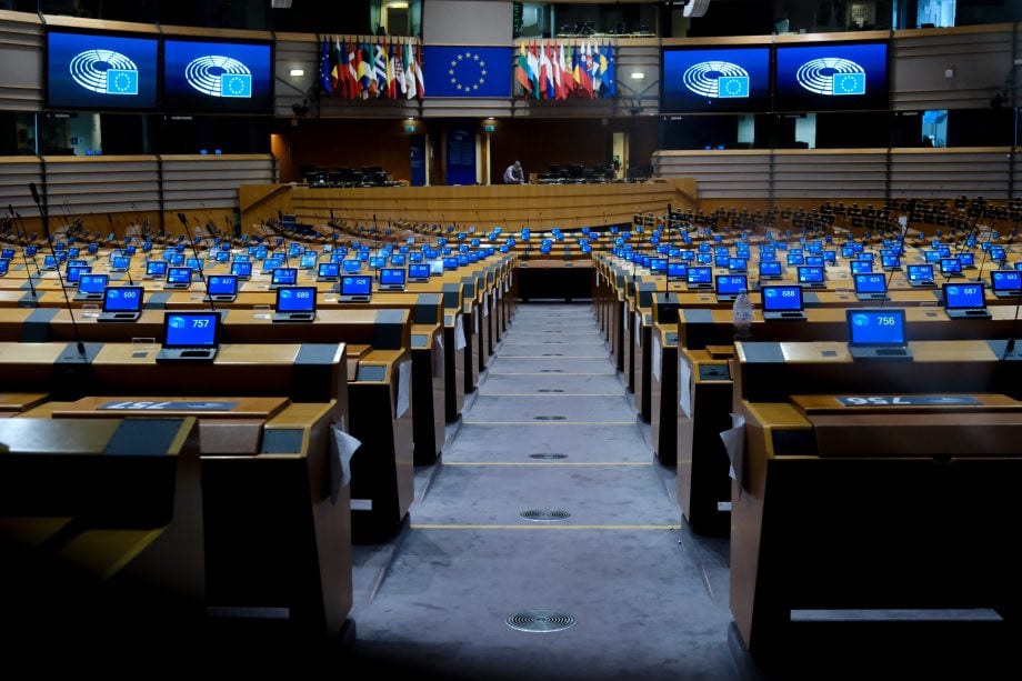 Das Bild zeigt den Plenarsaal des Europäischen Parlaments in Brüssel. Zu sehen sind die weitgehend leeren Sitzreihen der Abgeordneten. Auf mehreren Bildschirmen ist das Logo des Europäischen Parlaments vor blauem Hintergrund eingeblendet, im Zentrum hängen die EU-Flagge sowie die Flaggen der Mitgliedstaaten.