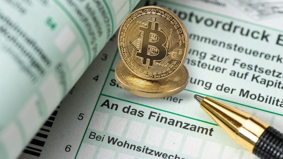 Man sieht einen Vordruck vom Finanzamt. Rechts davon liegt ein Kugelschreiber ab und mittig oben sind zwei Bitcoin-Münzen auf dem Formular positioniert.
